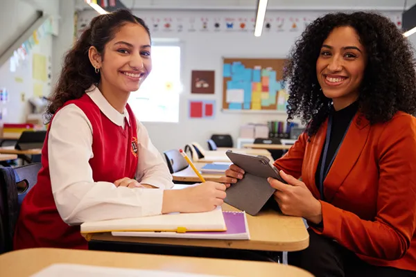  A teacher and student smiling in a classroom; the student is ready to write while the teacher references myON on a tablet